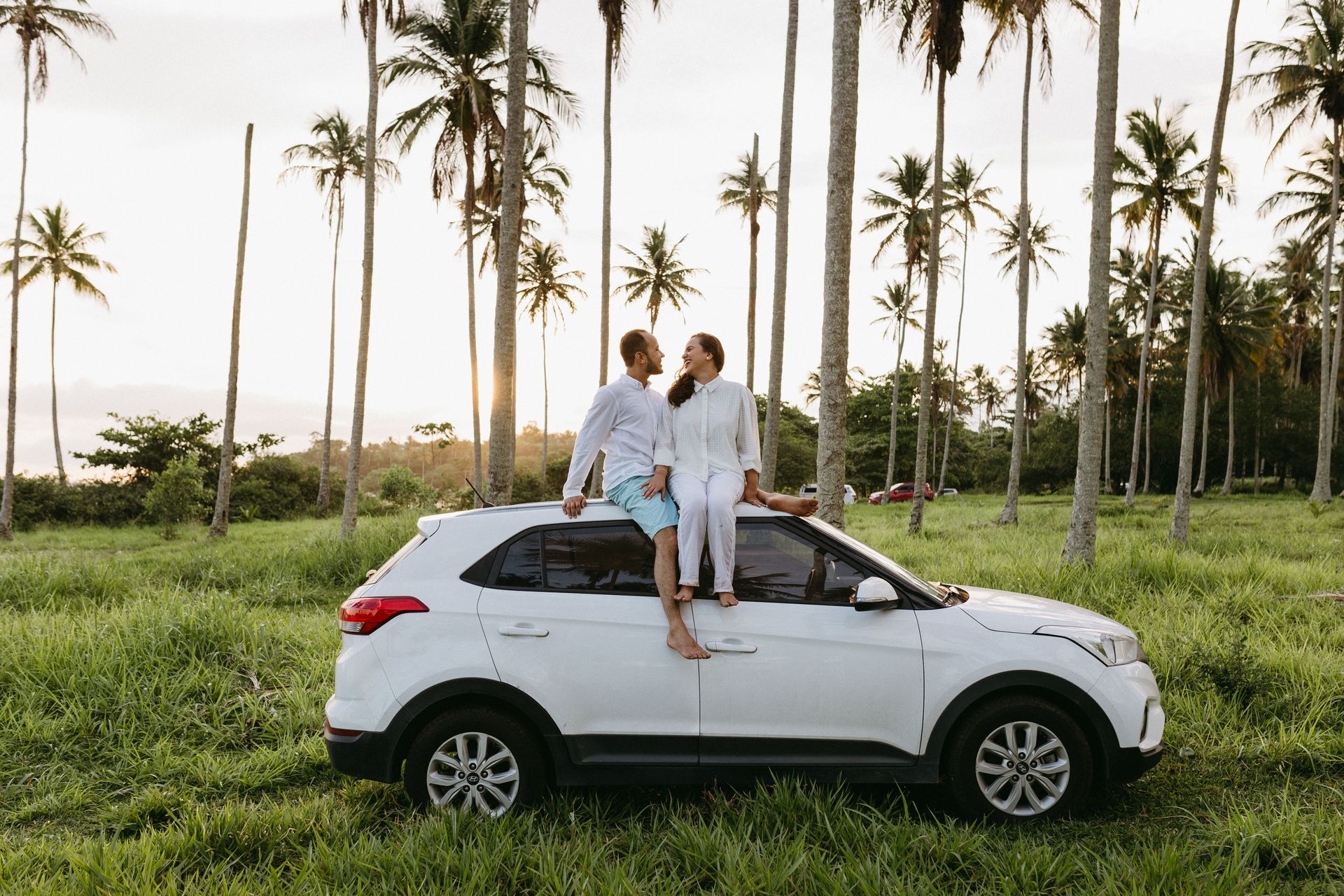 A romantic couple sits on a car roof amidst tropical palm trees at sunset, exuding happiness.